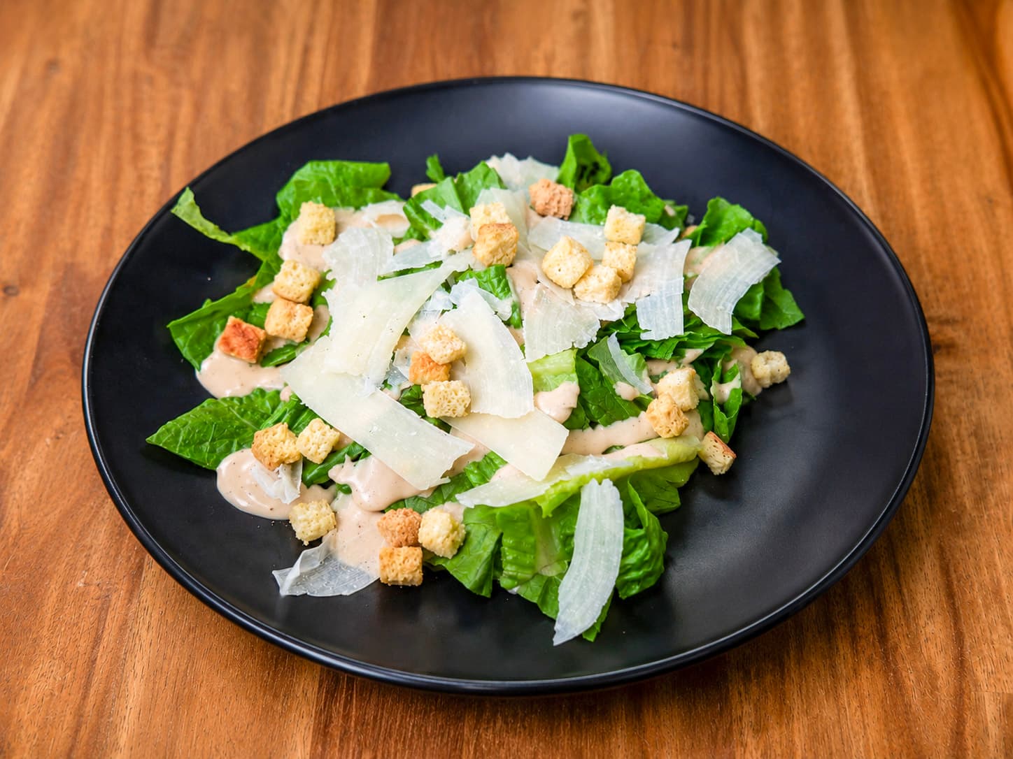Caesar salad with romaine, shaved parmesan, croutons, and caesar dressing on a black plate